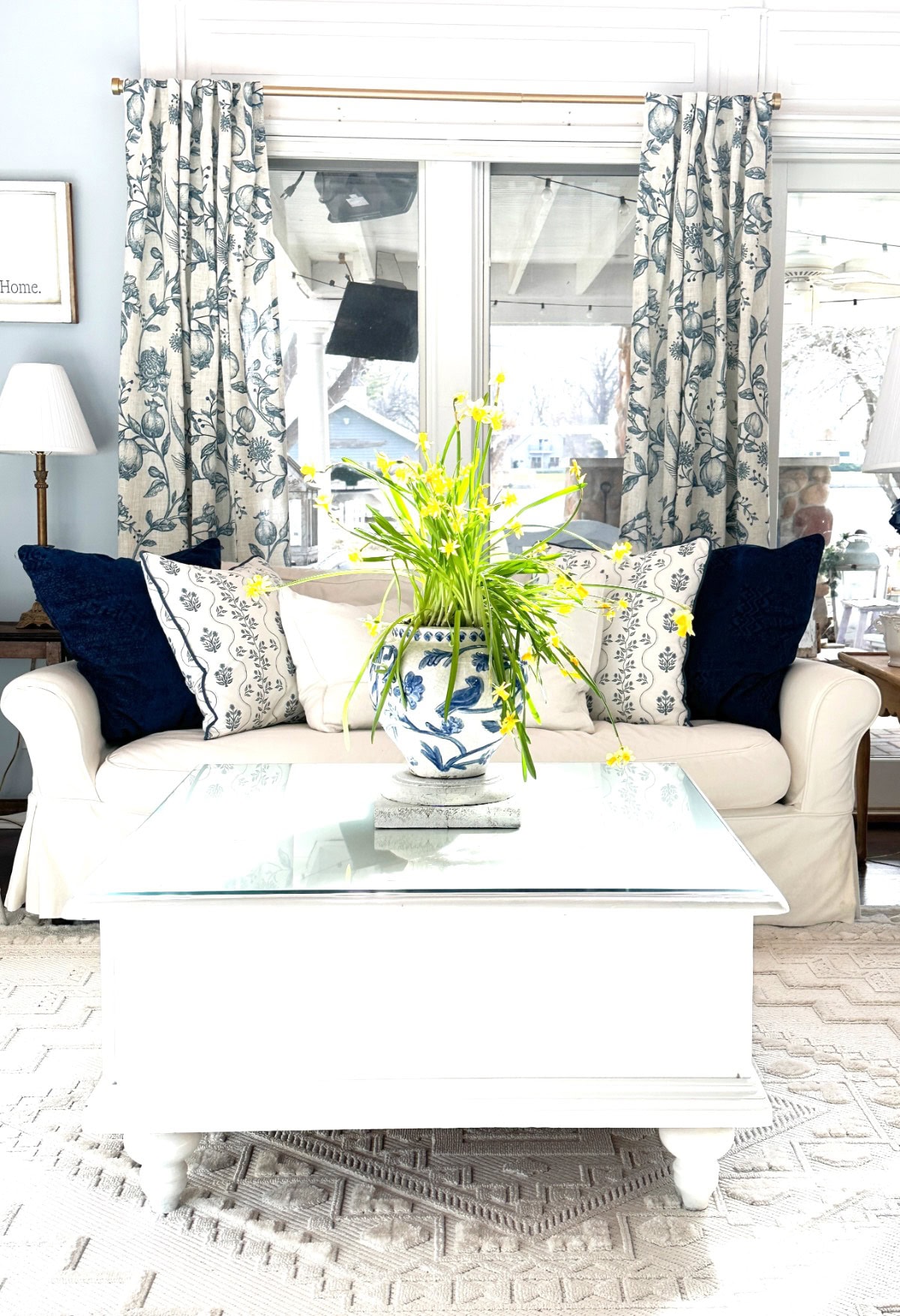 A bright living room exemplifies "Living Large in A Small House" with its white sofa adorned with blue and white patterned pillows. A glass coffee table holds a vase of yellow flowers, while floral curtains frame expansive windows, complemented by a stylish lamp on the left.