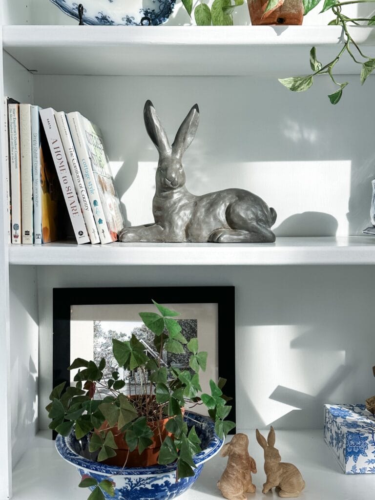 A shelf with a grey rabbit sculpture, books, and a plant in a patterned blue bowl showcases treasures from Saturday shopping. Below, a framed picture, small decorative rabbits, and a blue patterned box are displayed. Sunlight casts soft shadows across the scene.