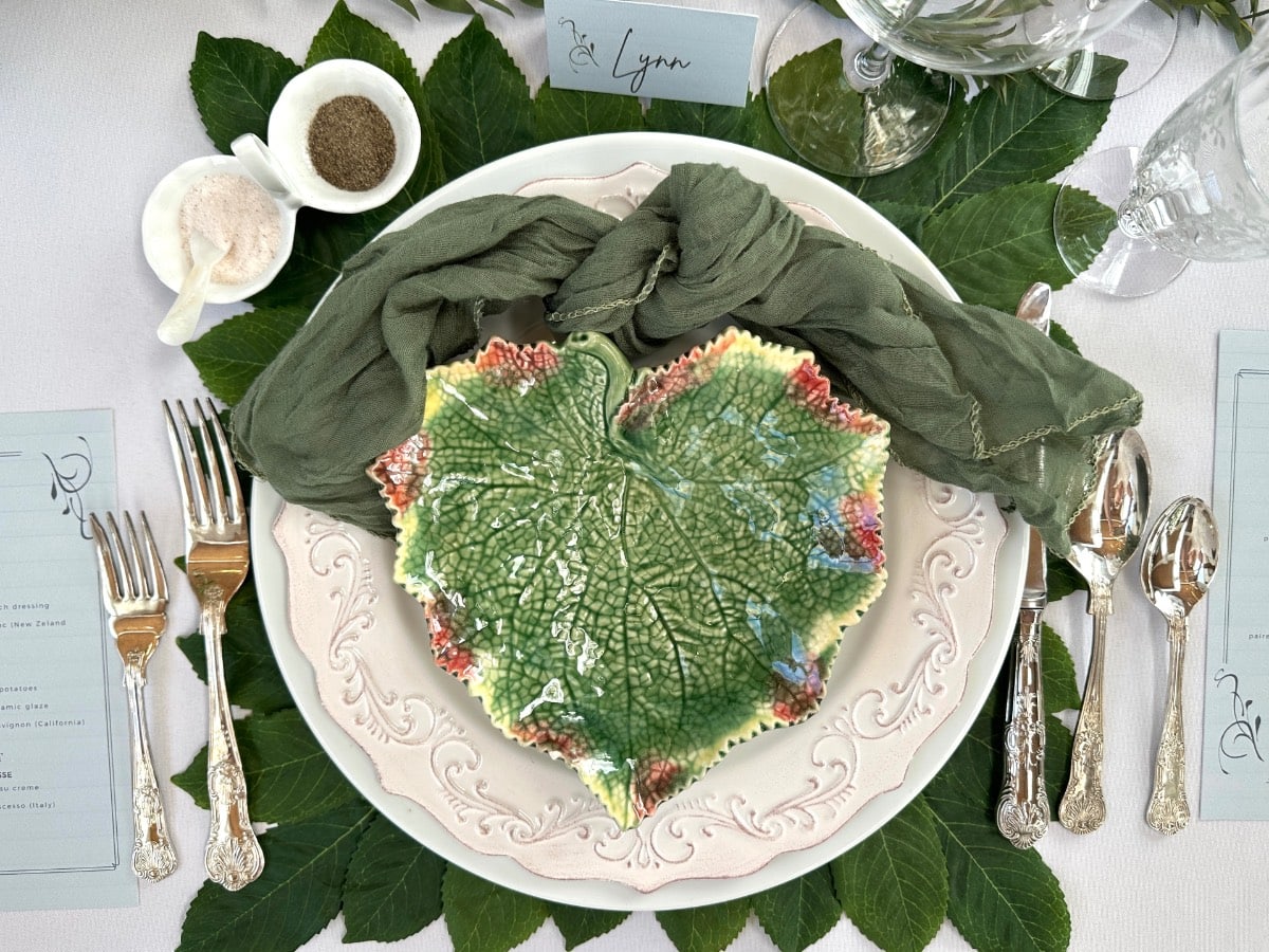 A detailed green leaf-shaped plate takes center stage on a white plate, surrounded by lush green leaves. Silverware is elegantly arranged, with glassware and menu cards adding charm—perfect for displaying Saturday shopping finds. A green cloth napkin is neatly folded above the plates.