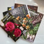 A collection of 2025 gardening catalogs is spread out. The catalogs feature colorful images, including pink and orange roses and leafy greens. Prominent titles include "Heirloom Roses" and "Johnny's Selected Seeds.