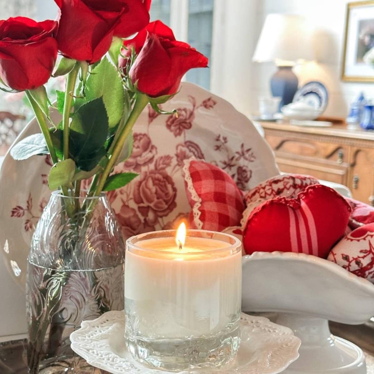 A lit candle in a glass holder sits on a white plate. Beside it is a wine bottle candle next to a vase of red roses, while a ceramic bowl cradles red and white patterned fabric hearts. The wooden sideboard in the background hosts decorative plates and a lamp.