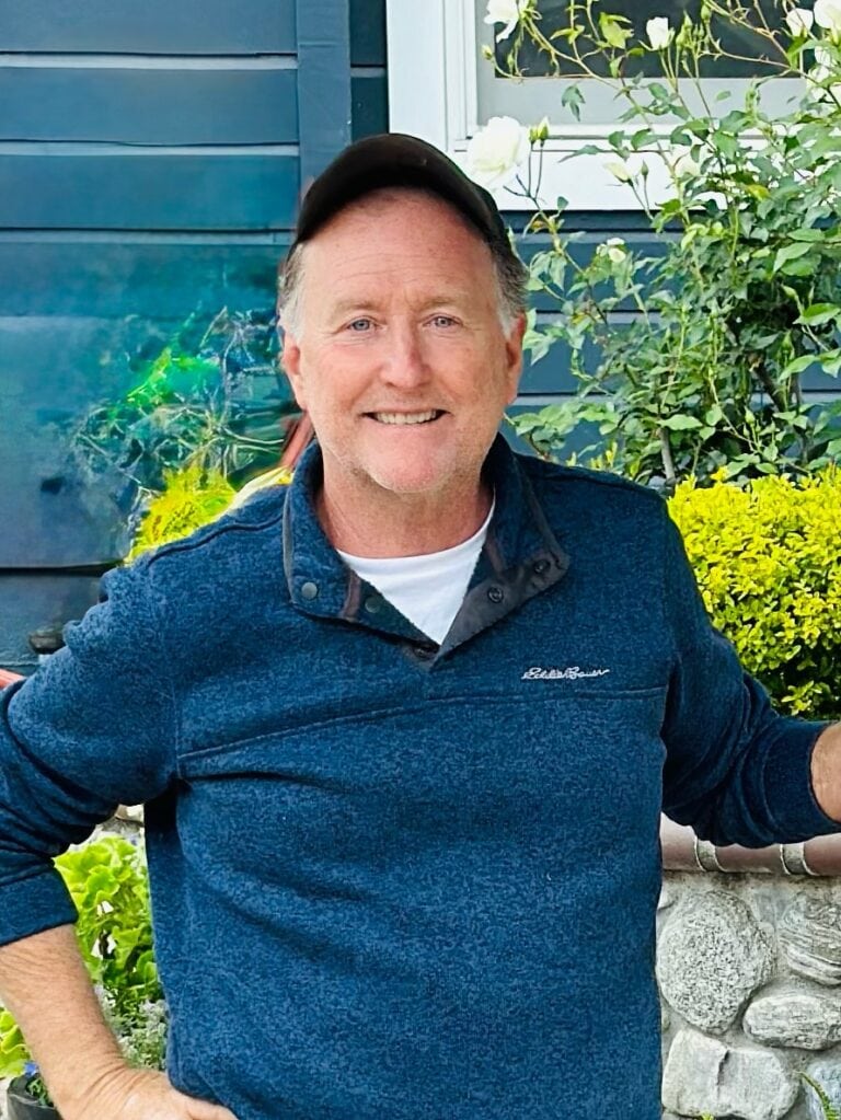 Mark Harris in a blue sweater and cap smiles while standing outdoors, possibly sharing tales from his latest podcast. Behind him are various plants and bushes against a dark blue wall. He rests one hand on a stone surface, exuding a relaxed confidence.