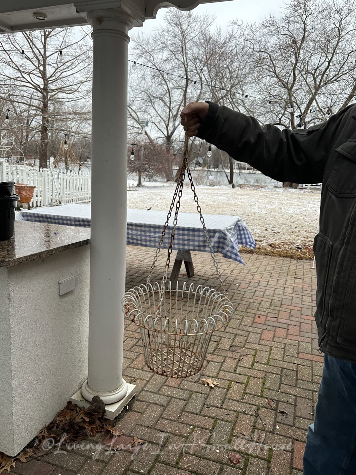 A person stands on a brick patio, holding an empty wire basket with chains. Nearby, a table adorned with a blue-checked tablecloth and a wine bottle repurposed as a candle holder adds charm to the snowy yard and leafless trees in the background.