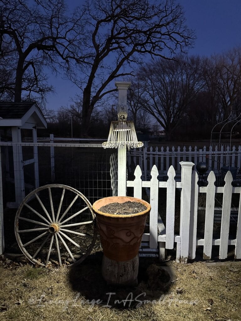 At dusk, a rustic garden scene reveals a wine bottle candle glowing atop a large terracotta pot on a tree stump, casting flickering shadows. Behind it, an upside-down wire basket sits mounted on the white picket fence beside a wooden wagon wheel. Bare trees loom in the background.