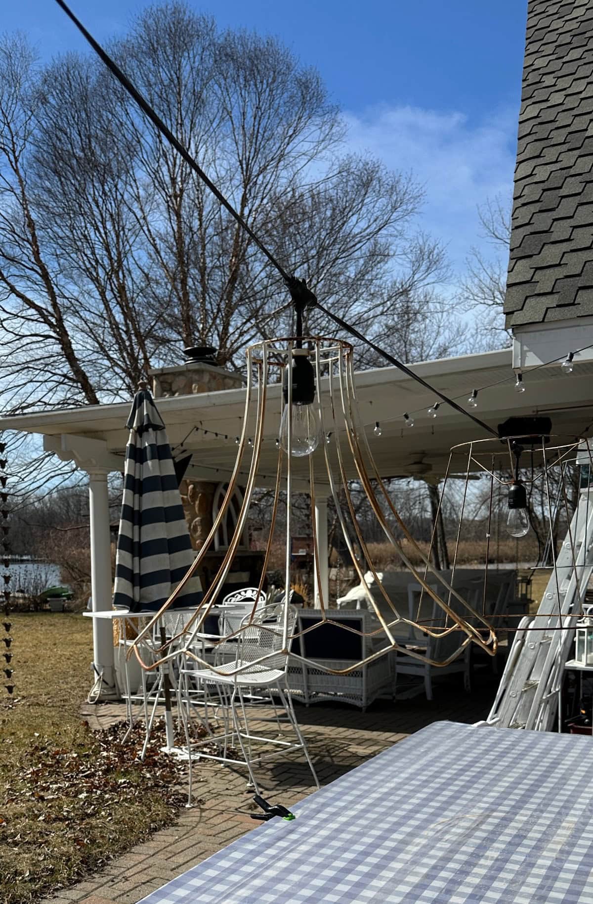 A patio with a checkered tablecloth on the table, where a large light fixture hangs from a string, accompanied by a striped umbrella near white outdoor furniture under a pergola. A wine bottle candle adds charm, while bare trees and a partly cloudy sky complete the backdrop.