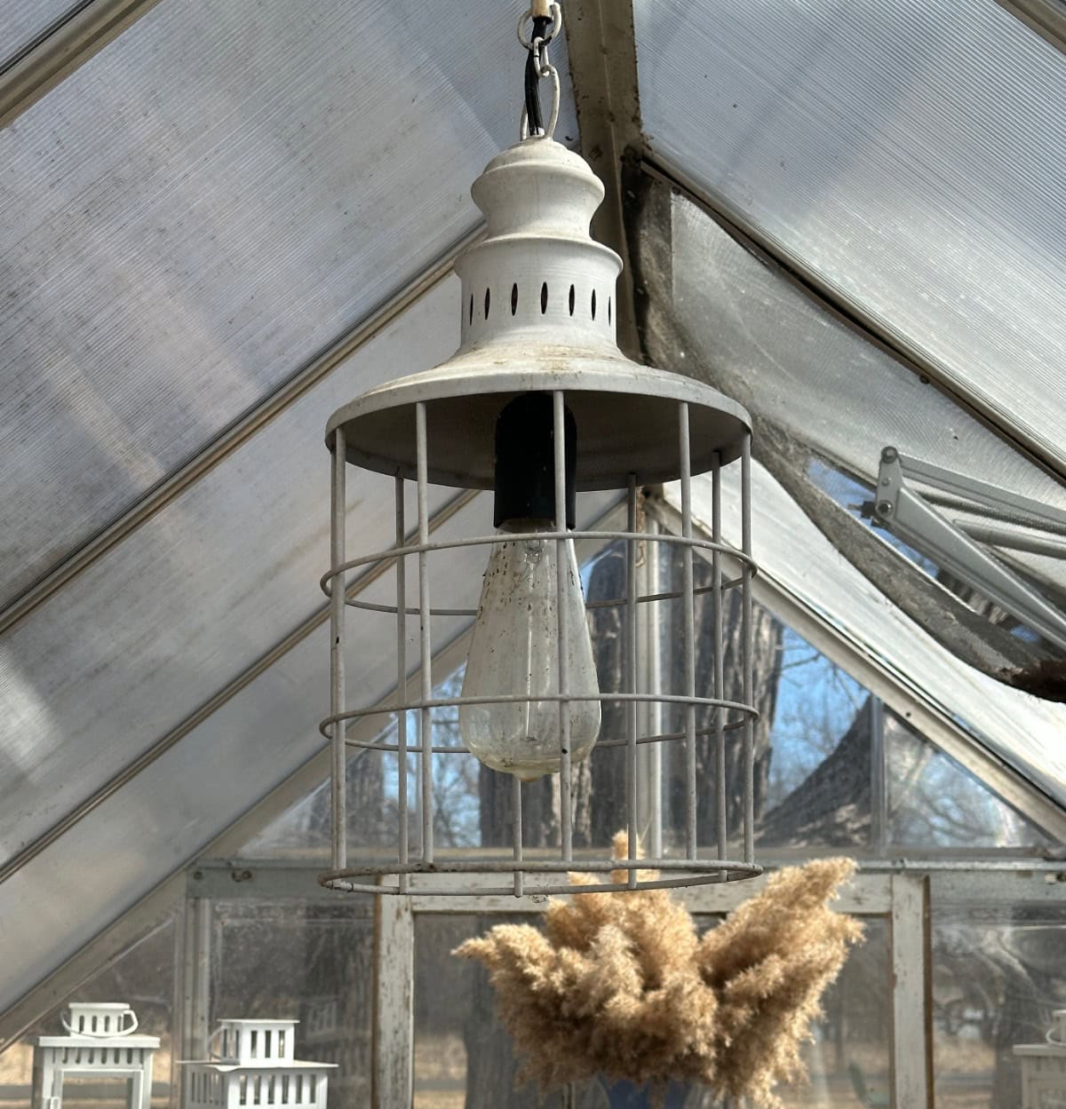 A rustic metal lantern-style light fixture, reminiscent of a wine bottle candle ambiance, hangs from the ceiling of a greenhouse. Dried plants are visible below the light as sunlight filters through the glass roof and windows, illuminating the space.