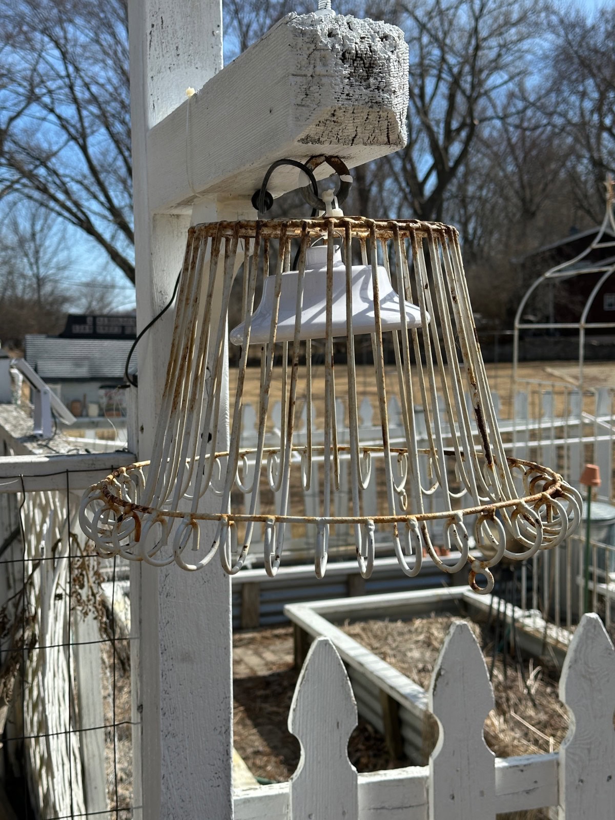 A rustic light fixture crafted from an upside-down metal lampshade frame features a wine bottle candle. Attached to a white wooden post, it hangs near a white picket fence with a garden in the background, framed by trees beneath a clear sky.