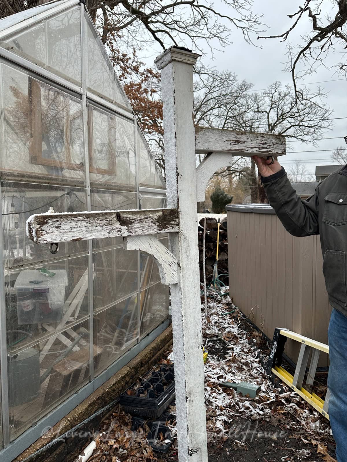 A person holds a weathered, white wooden structure resembling a tall T-shaped coat rack outside next to a greenhouse. Nearby, a wine bottle candle flickers among leaves and a dusting of snow. In the background, a shed and some scattered tools complete the rustic scene.