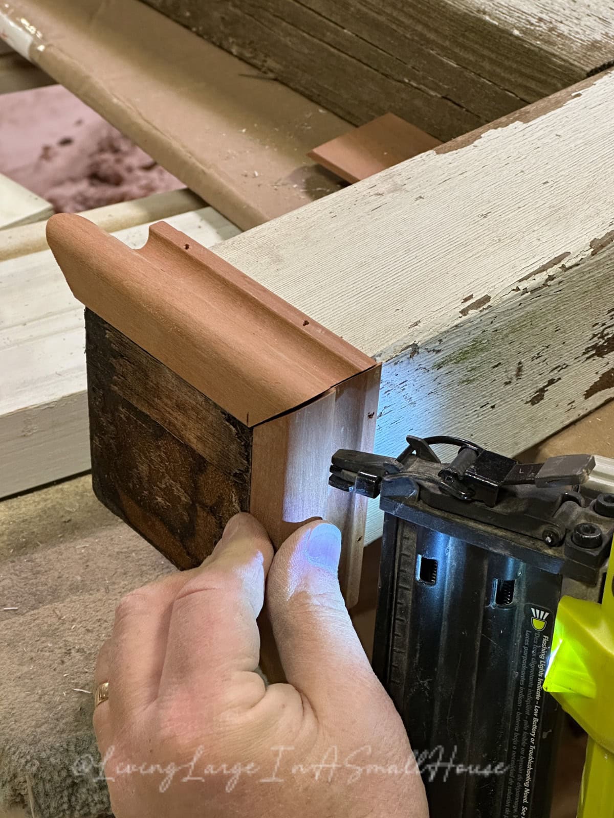 A person skillfully uses an electric nail gun to attach a small piece of wood to larger wooden structures. In the background, hues of wood mingle with the soft glow of a wine bottle candle, adding warmth to the scene captured in close-up.