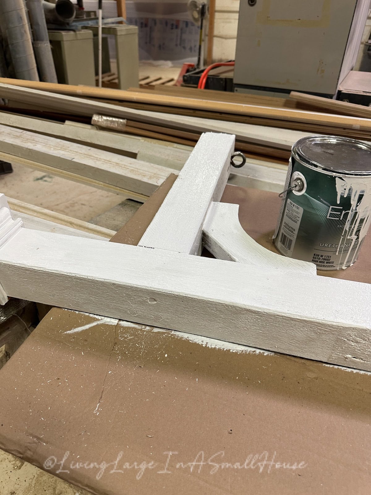 A carpentry workshop features wooden pieces painted white on a workbench, accompanied by a can of paint and brushes. A wine bottle candle flickers gently nearby, casting soft light over the paint-splattered floor and various tools and equipment in the background.