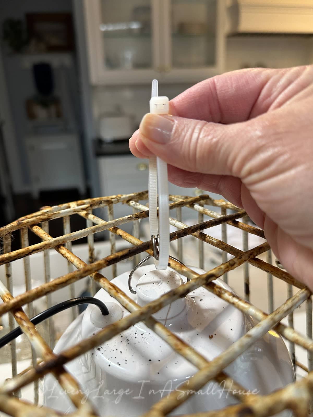 A hand is securing a white cable tie to the chain of a rusty metal birdcage, inside which a white object resembles a light fixture or perhaps the glow from a wine bottle candle. The background reveals hints of a blurred kitchen setting.