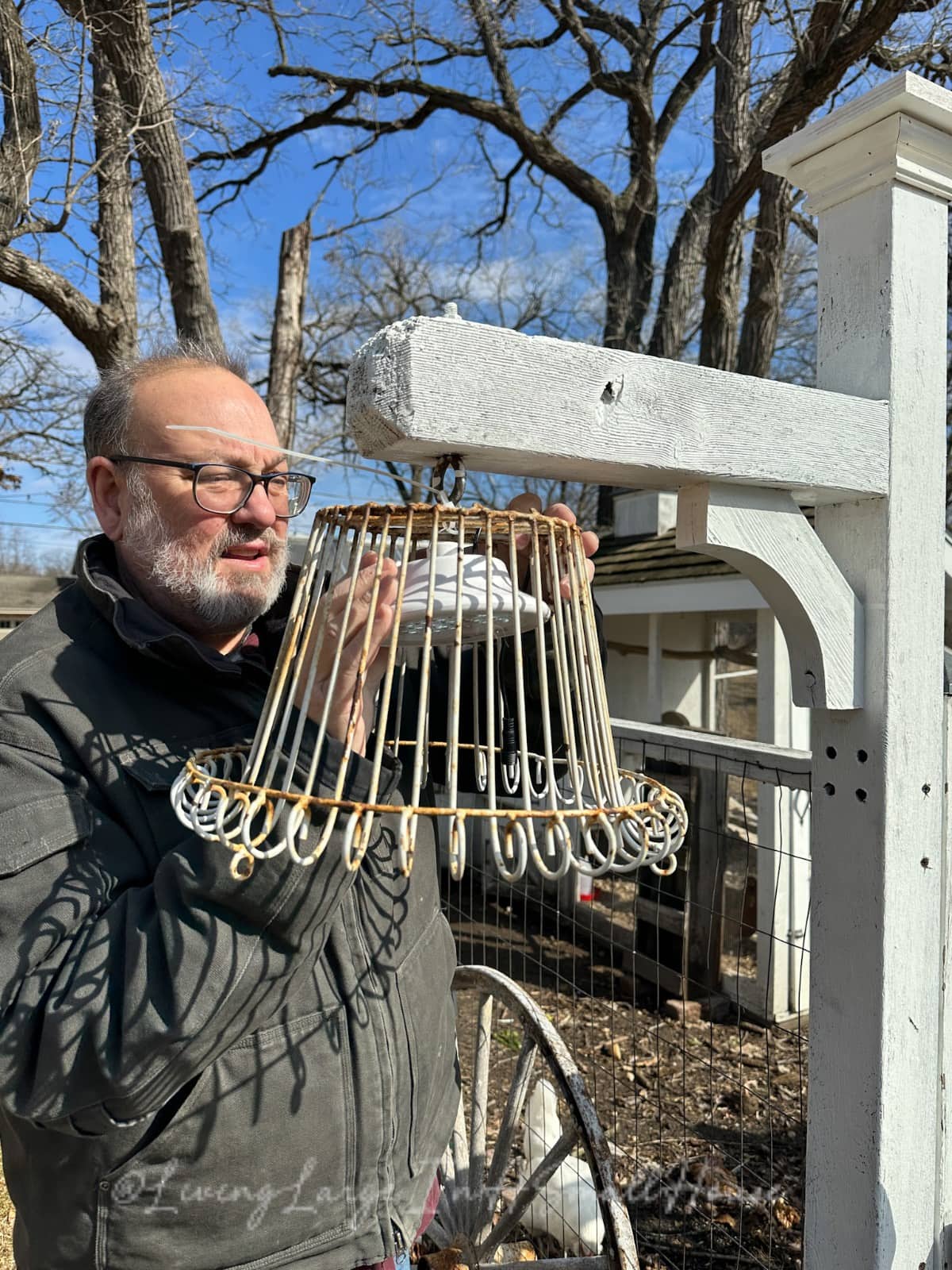 A person wearing glasses inspects a metal cage-like structure, reminiscent of an oversized wine bottle holder, hanging from a wooden beam amidst leafless trees. The person holds the structure with one hand as if contemplating its transformation into a unique candle holder.