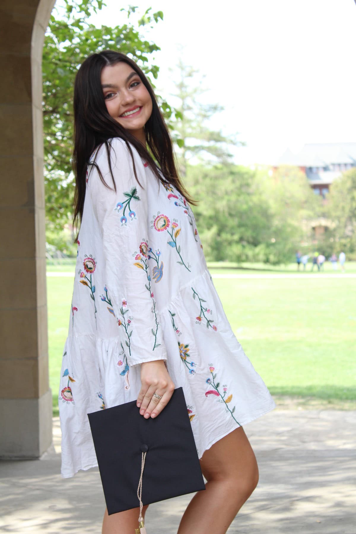 Smiling woman in a white, floral dress holds a graduation cap. She stands near an archway with greenery and a field visible in the background, conveying a celebratory mood.