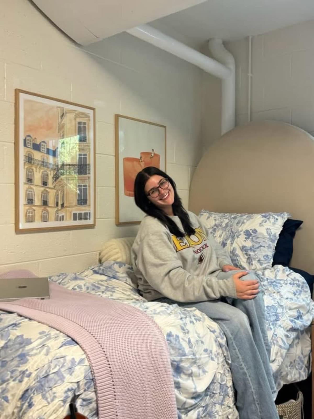 college-life-12 | Living Large In A Small House, LLC A person with glasses smiles while sitting on a bed in a cozy room. The bed has floral bedding and a pink blanket. Two framed pictures hang on the wall. A laptop rests on the bed beside them.
