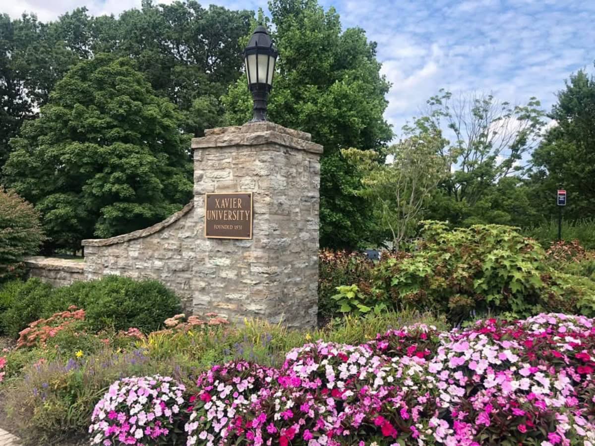 college-life-17 | Living Large In A Small House, LLC Stone entrance sign for Xavier University, established 1831, surrounded by vibrant pink and purple flowers. Mature trees and a cloudy blue sky form the picturesque backdrop.
