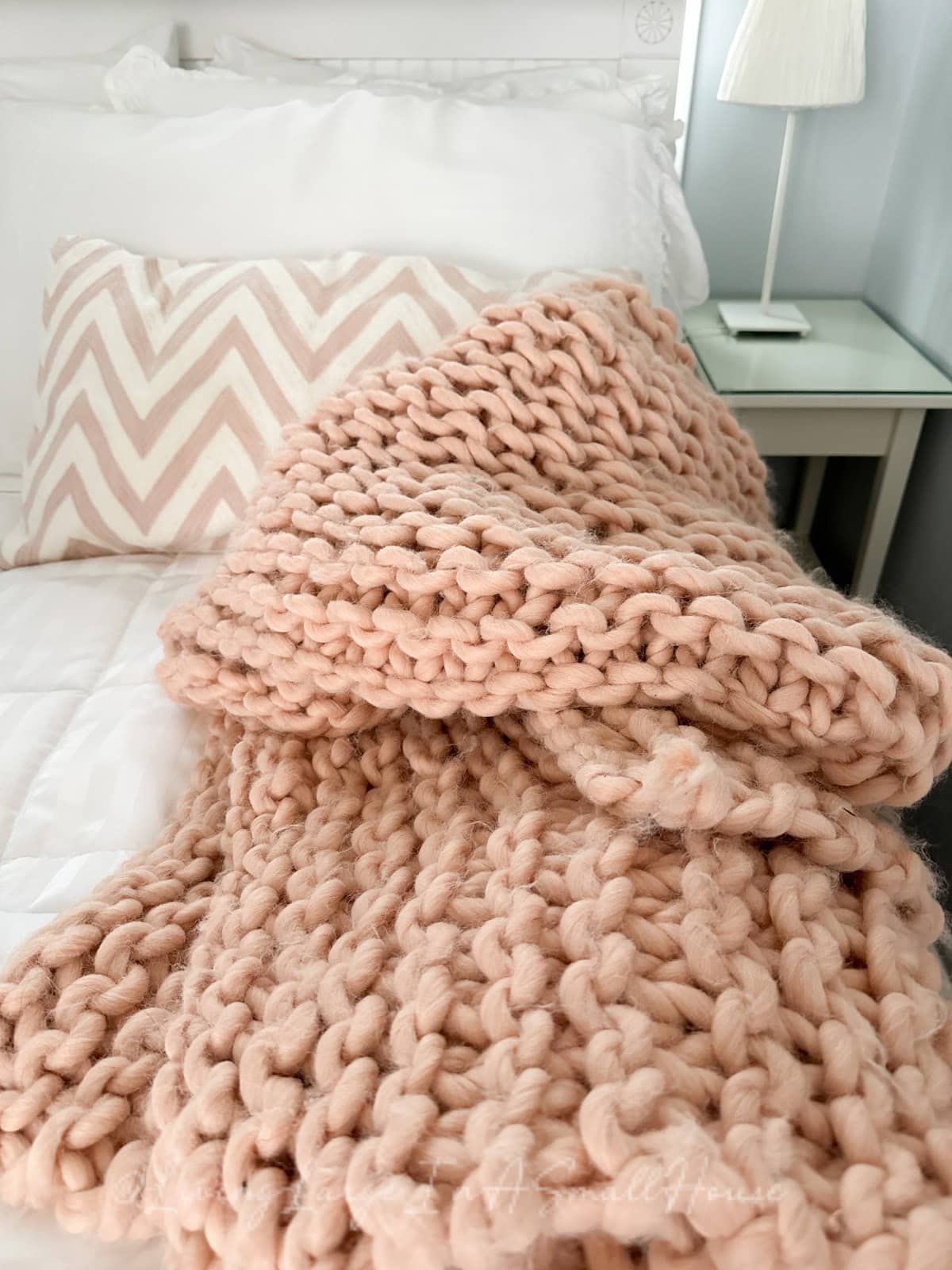 A bedroom scene with a bed featuring a chunky knit pink blanket draped over it. The bed has white sheets and a white pillow with a chevron-striped cover. A white nightstand and lamp are beside the bed.