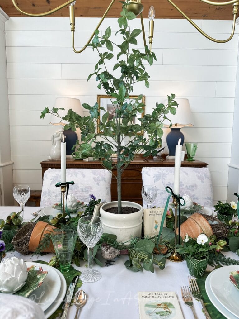 A dining table is elegantly set with green foliage, potted plant centerpiece, white candles, and garden-themed decor. Plates and glassware are arranged neatly, with a book titled "The Tale of Mrs. Tiggy-Winkle" visible.