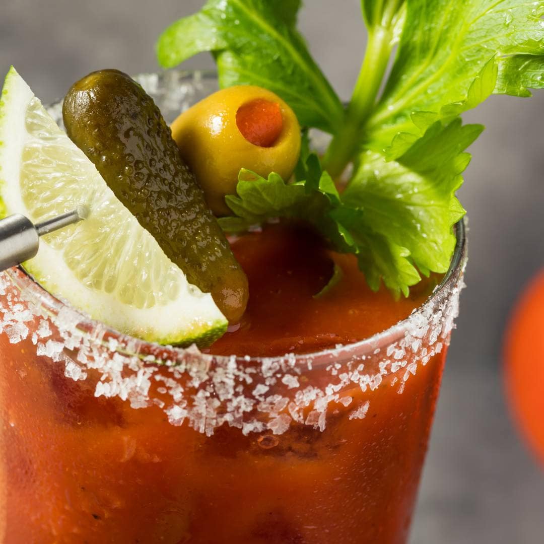 A close-up of a Bloody Mary cocktail with a salted rim, garnished with a lime wedge, pickle, green olive, and celery stalk—perfect for sipping after Saturday shopping.