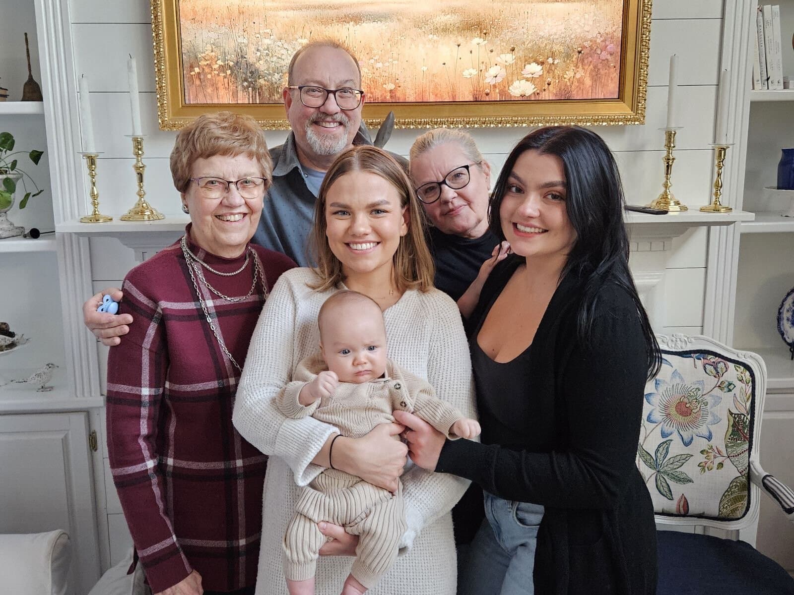 About six people, including a baby, stand close together and smile in a warmly lit living room. The adults surround the baby, held by a woman in the center. A colorful painting hangs on the wall behind them.