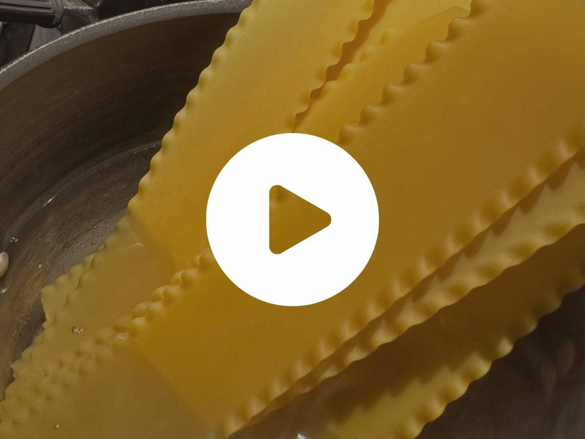 Close-up of uncooked lasagna noodles partially submerged in water in a pot, with a play button overlayed on the image.