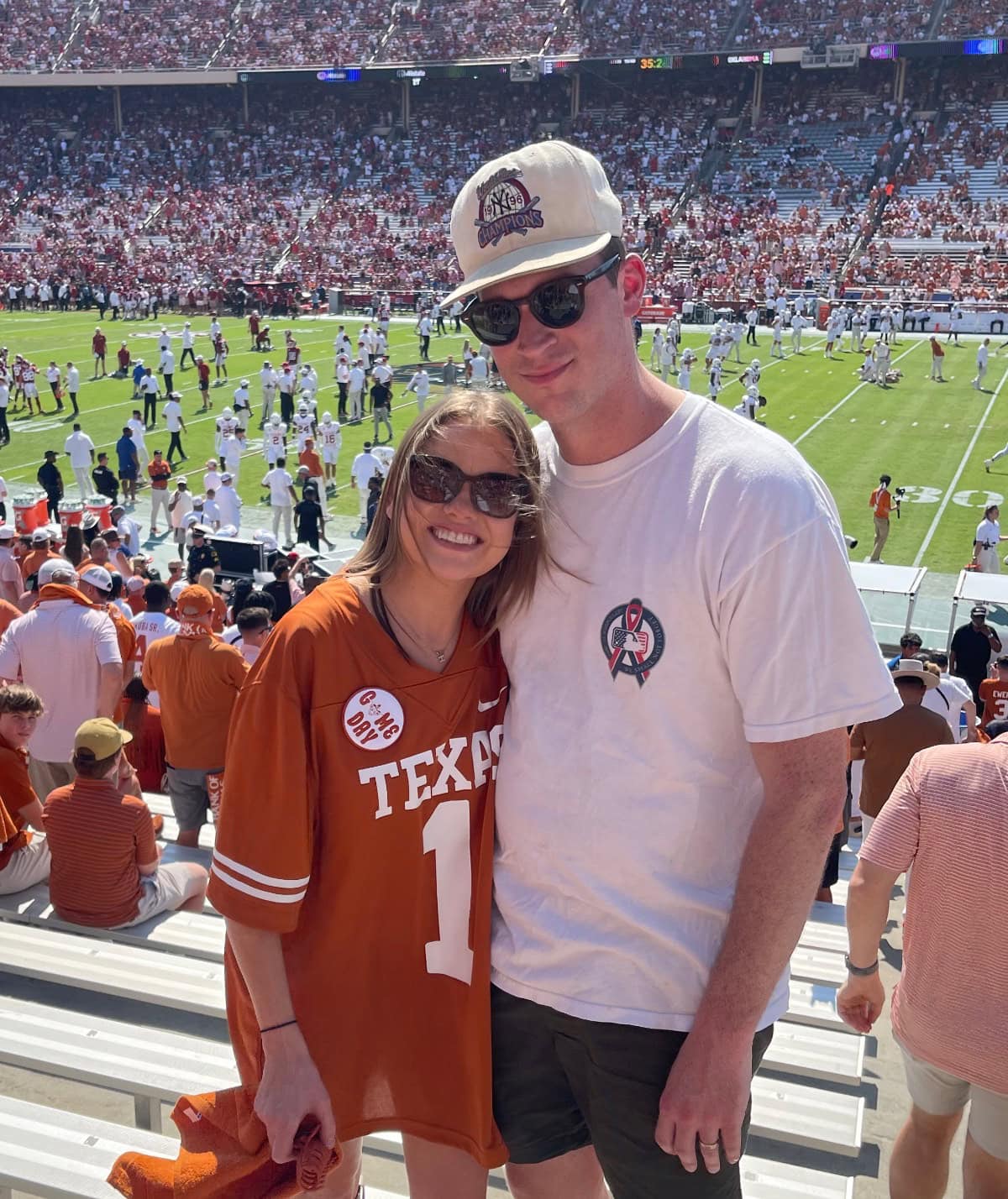 About: A smiling couple poses in a stadium with a football game in progress behind them. The woman wears a burnt orange Texas jersey and sunglasses; the man sports a cap, sunglasses, and white shirt. The stands are packed with fans.