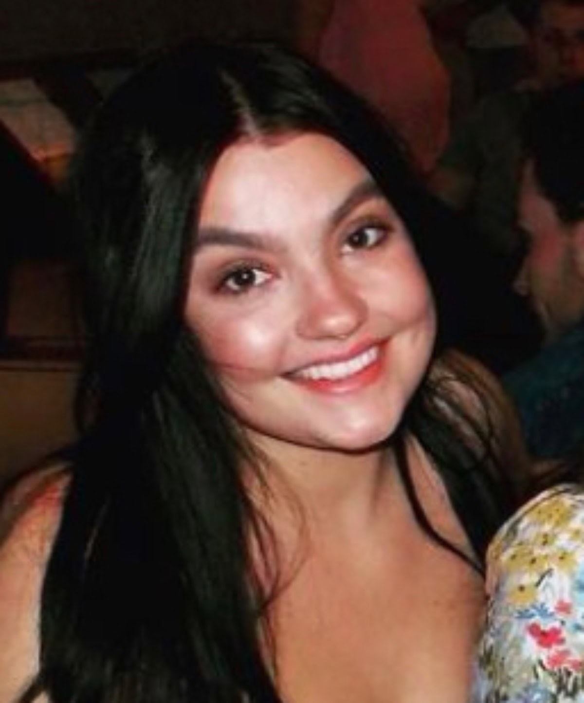 A woman with long dark hair and a floral top is smiling at the camera in a warmly lit indoor setting.