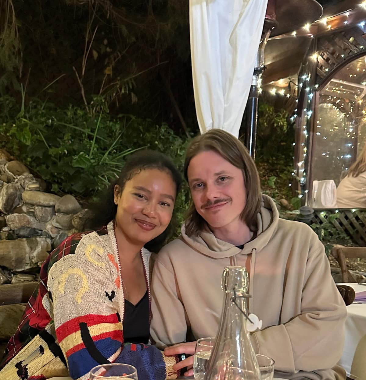 A smiling couple sits closely together at an outdoor restaurant table at night, surrounded by greenery and fairy lights. It's all about enjoying a relaxed and happy evening, with drinks and a water bottle on the table in front of them.
