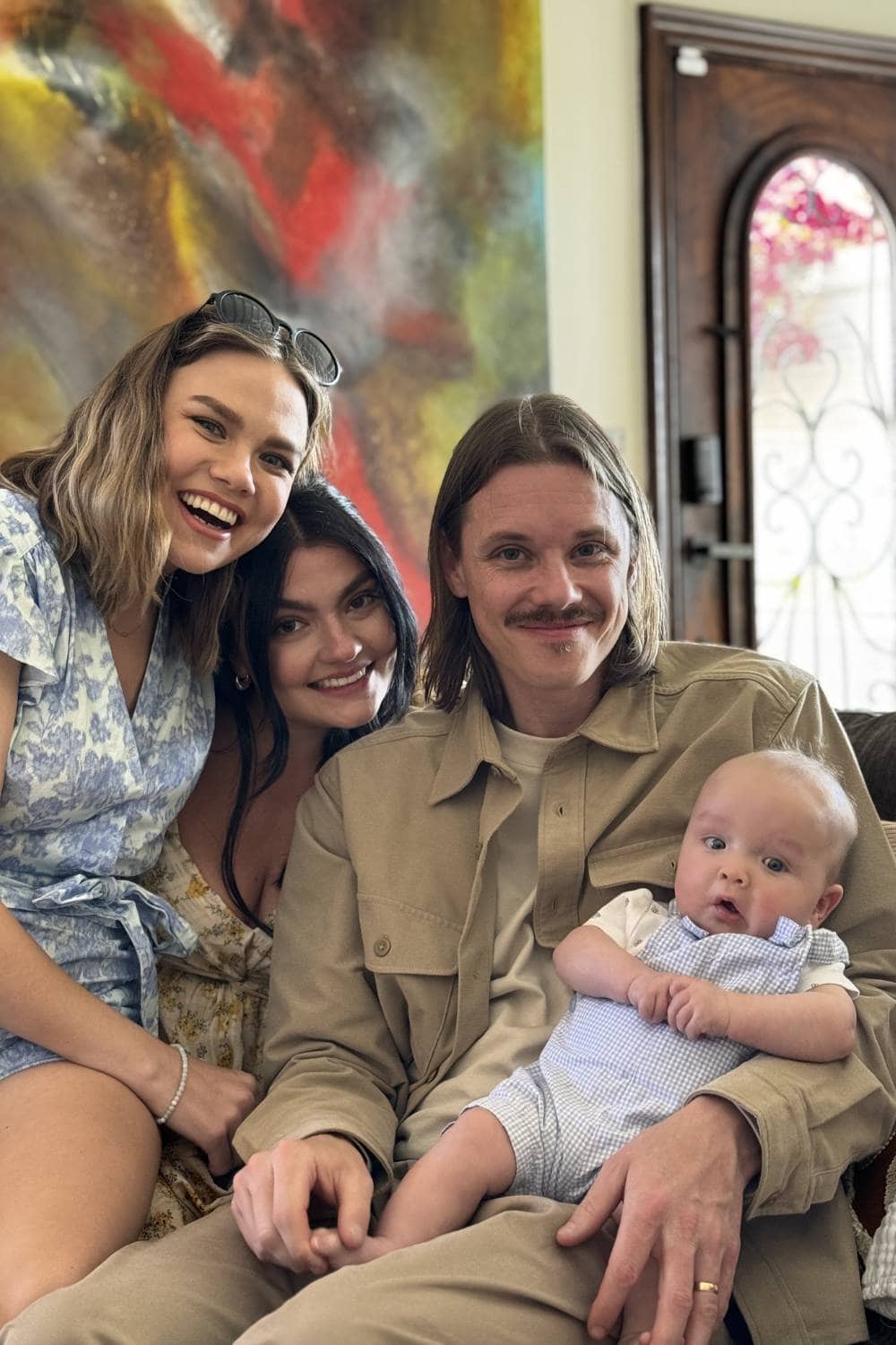 Three adults and a baby sit together indoors, smiling at the camera. The man holding the baby has a mustache, and two women are closely seated beside him. A colorful painting and a wooden door are visible in the background, adding warmth to this about family moment.