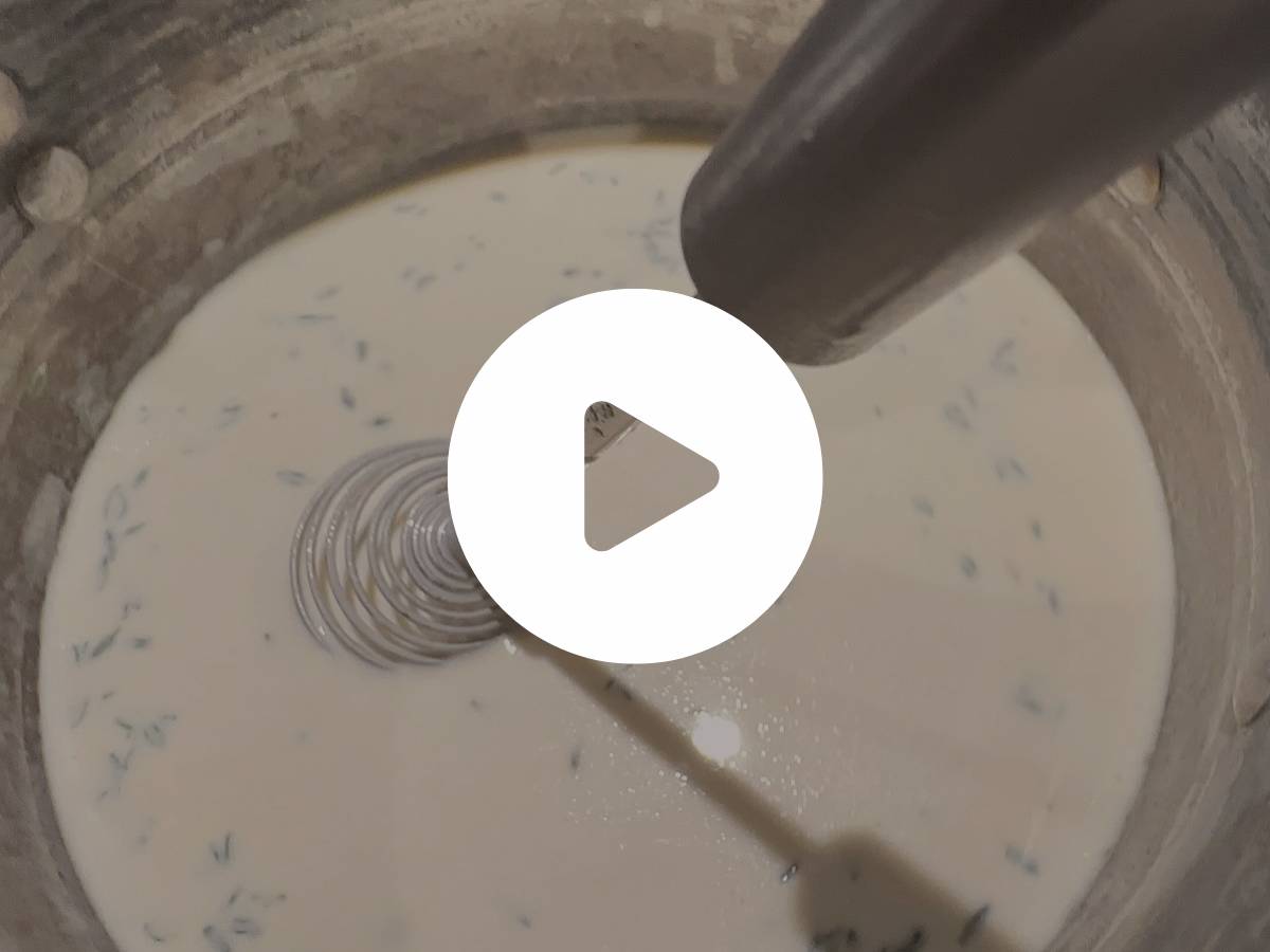 A metal whisk and blender are mixing a creamy white liquid with small green herbs in a bowl. The scene is viewed from above, and a play button is overlayed on the image, suggesting it's a video.