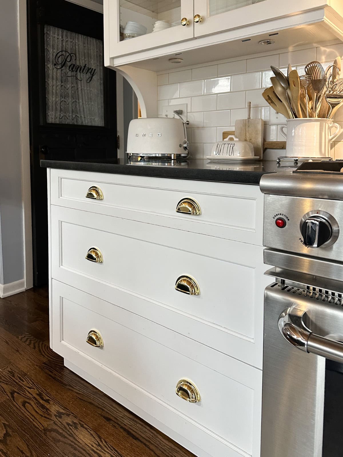 A kitchen with white cabinets and gold handles, a black countertop with a toaster, butter dish, and various utensils in a jar. The stove is partially visible on the right, and a door with "Pantry" written on it is in the background.