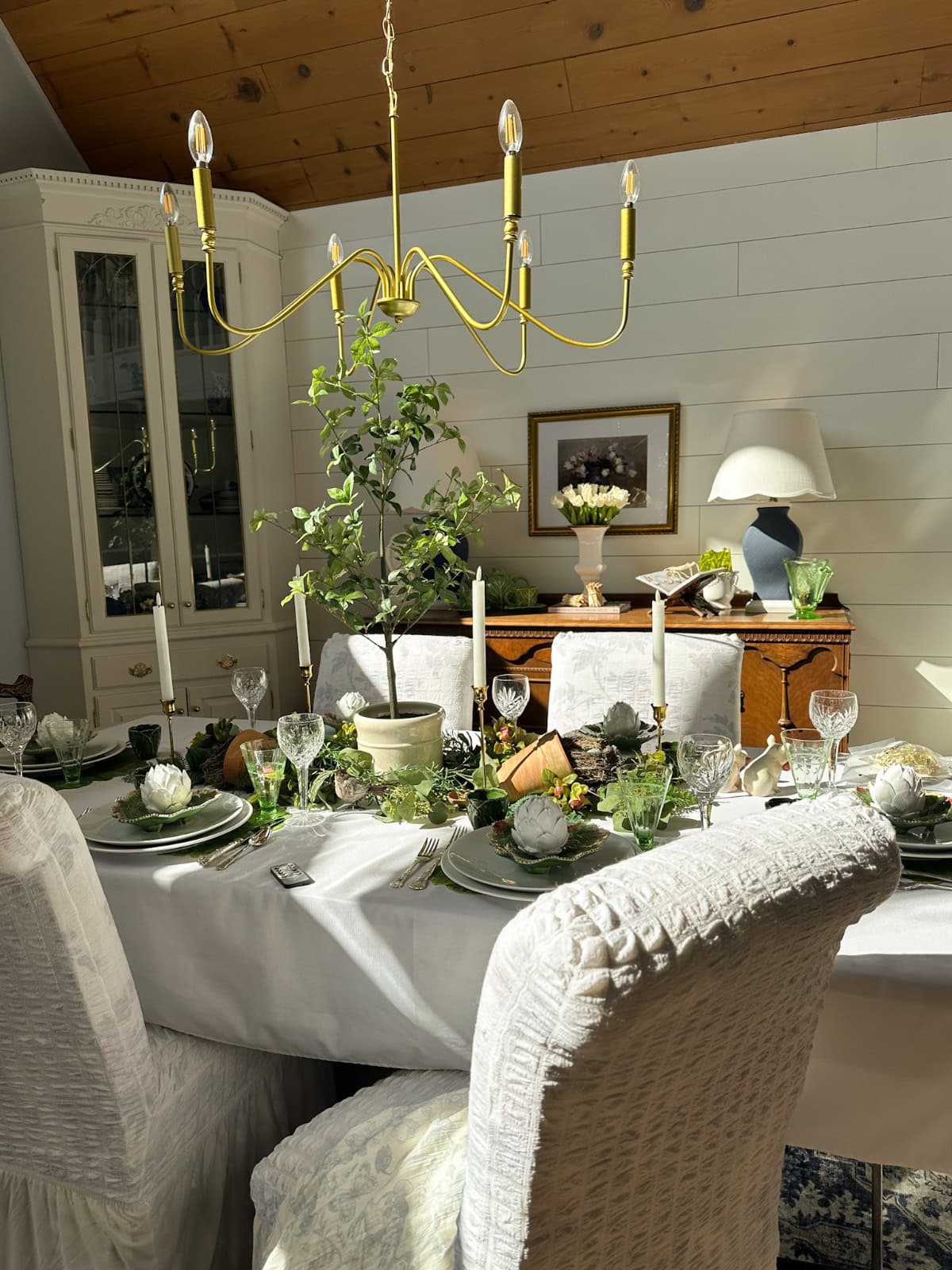 A dining table is elegantly set with white and green floral arrangements and candles. Crystal glassware and a chandelier enhance the setting. White chairs are covered in textured fabric. A wooden cabinet and a potted plant are in the background.
