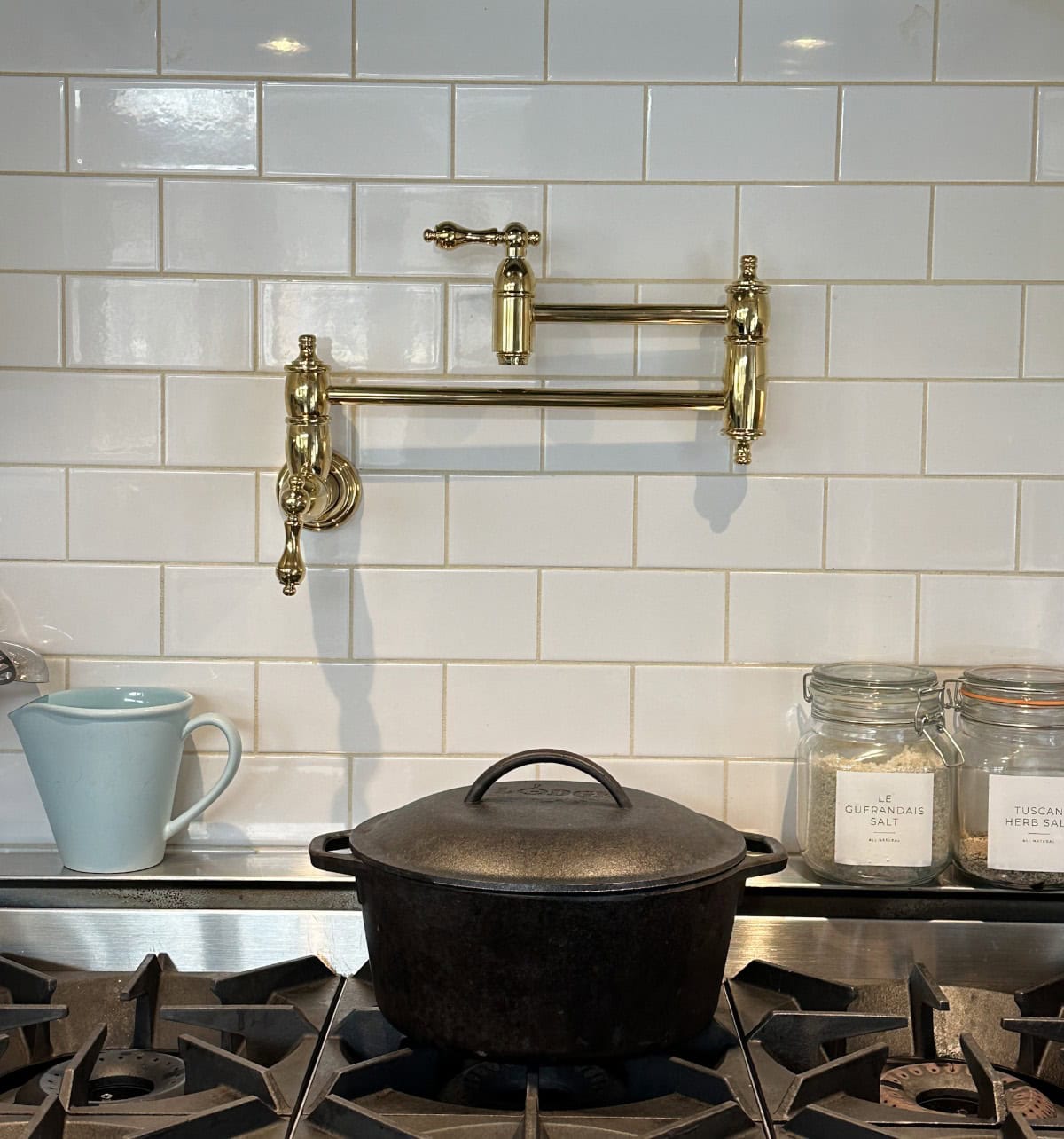 A cast iron pot sits on a stove. Behind it is a white subway tile backsplash with a brass pot filler faucet. To the right are jars of salt and spices, and to the left is a light blue pitcher.