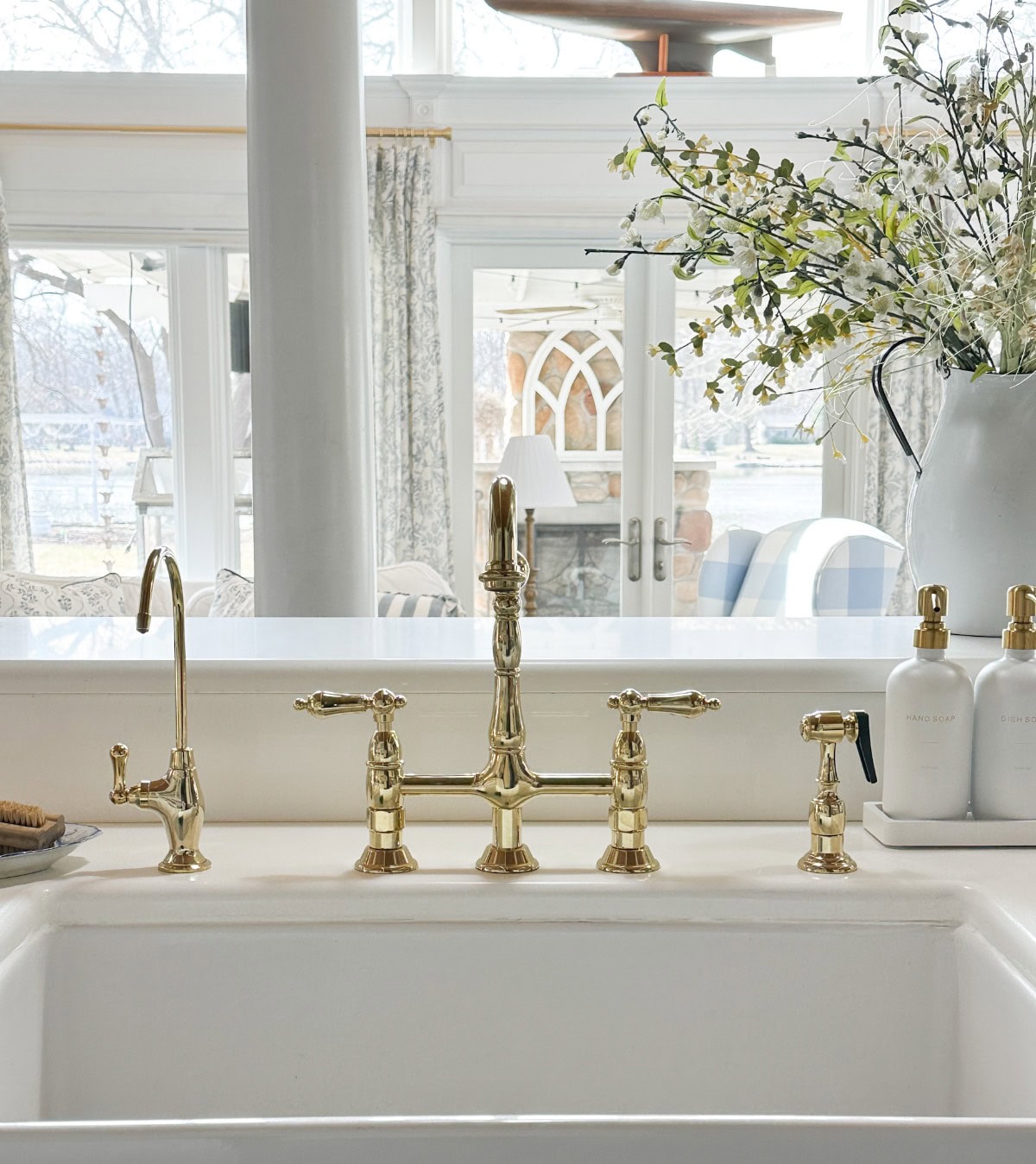 A bright kitchen with a white countertop featuring a gold faucet and soap dispensers. A large bouquet of flowers is placed on the counter. In the background, large windows and a glass door offer a view of a patio with outdoor seating.