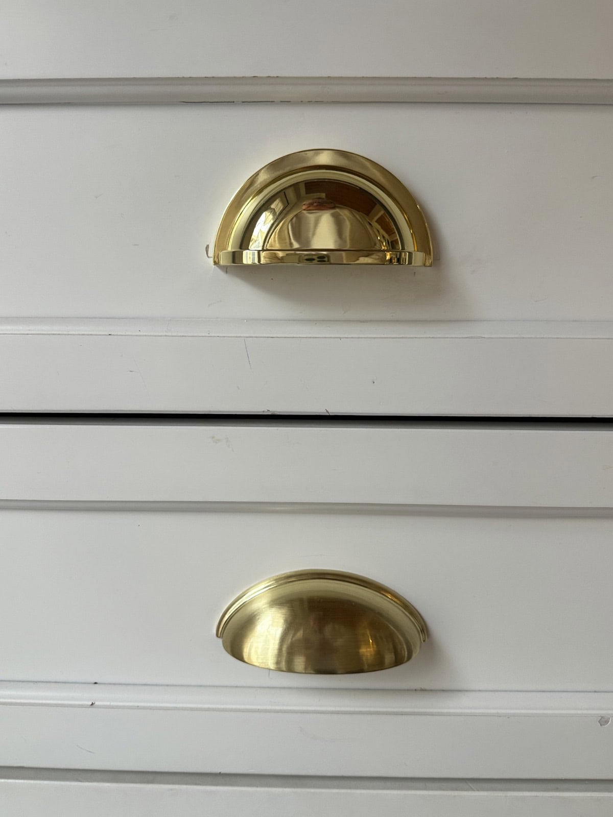 Two white drawers with brass cup handles. The drawers are close together, and the handles have a shiny, reflective surface. The image focuses on the handles, highlighting their metallic finish against the white background.