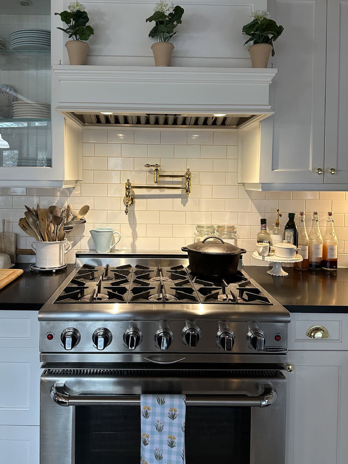 A modern kitchen stove with a stainless steel gas range, large black pot, and tea towel. White cabinets and backsplash, wooden utensils in a holder, condiments, and decorative plants on a shelf above. Subtle lighting creates a homely atmosphere.