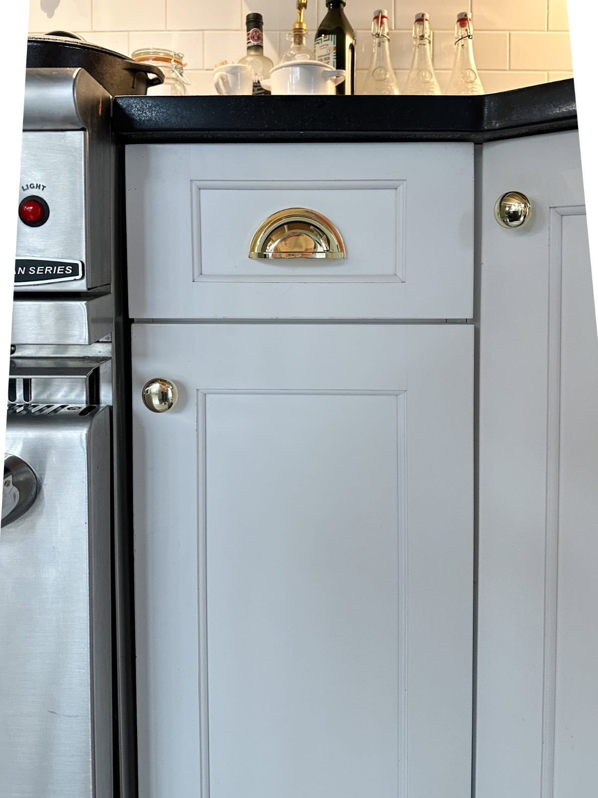 A kitchen cabinet with white panels and brass handles is next to a stainless steel appliance. On the black countertop above, there are bottles, a mug, and other small kitchen items against a white tile backsplash.