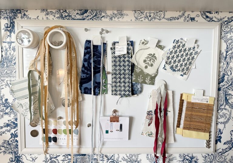 The whiteboard, reminiscent of a Saturday shopping spree, showcases an assortment of fabric swatches in various patterns and colors, ribbons, cutouts, and a color palette card. The background displays elegant blue and white toile-patterned wallpaper.