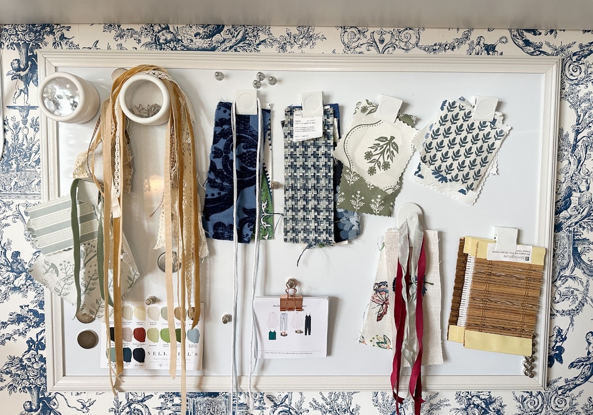 The whiteboard, reminiscent of a Saturday shopping spree, showcases an assortment of fabric swatches in various patterns and colors, ribbons, cutouts, and a color palette card. The background displays elegant blue and white toile-patterned wallpaper.