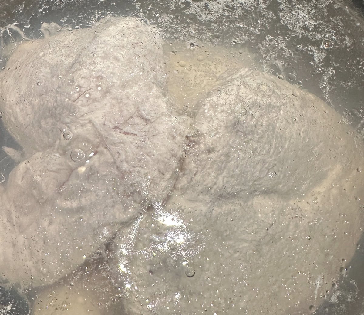 Close-up of raw chicken pieces submerged in water, appearing pale and textured. The surface is glistening with bubbles and reflections from the liquid.