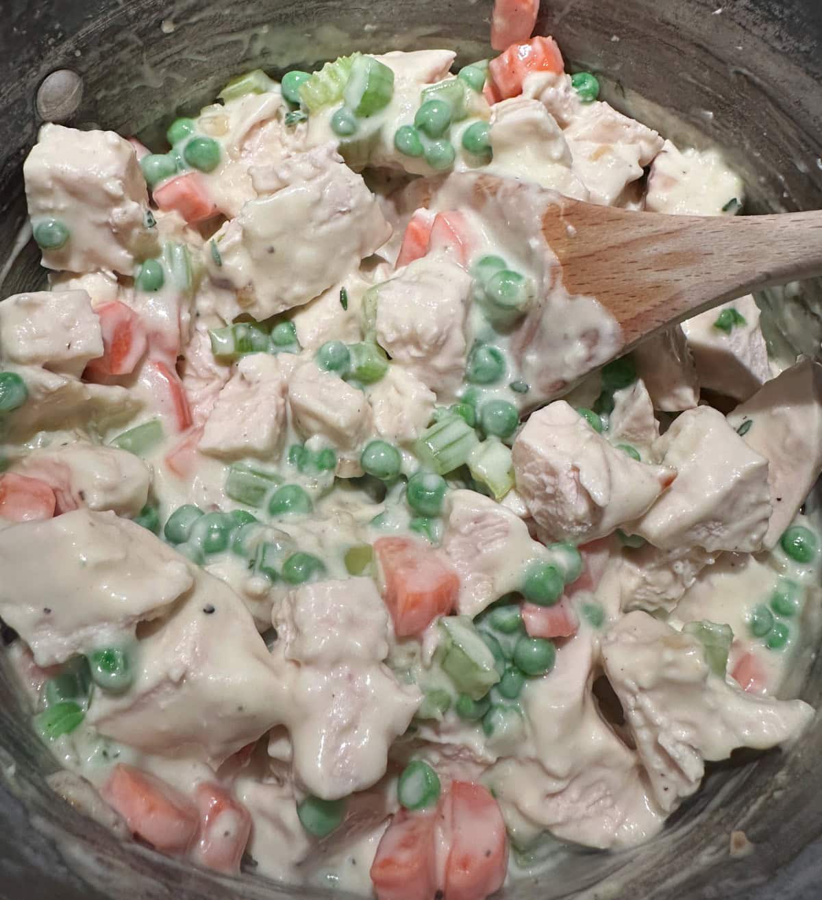 A creamy mixture of diced chicken, peas, carrots, and celery is being stirred with a wooden spoon in a pot. The ingredients are coated in a thick white sauce.