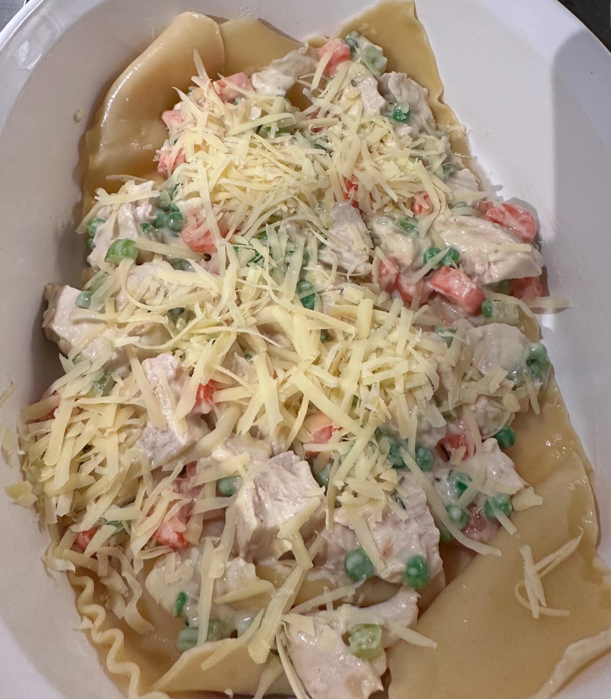 A dish of creamy pasta with chunks of chicken, peas, and diced carrots, topped with shredded cheese, in a white oval dish.
