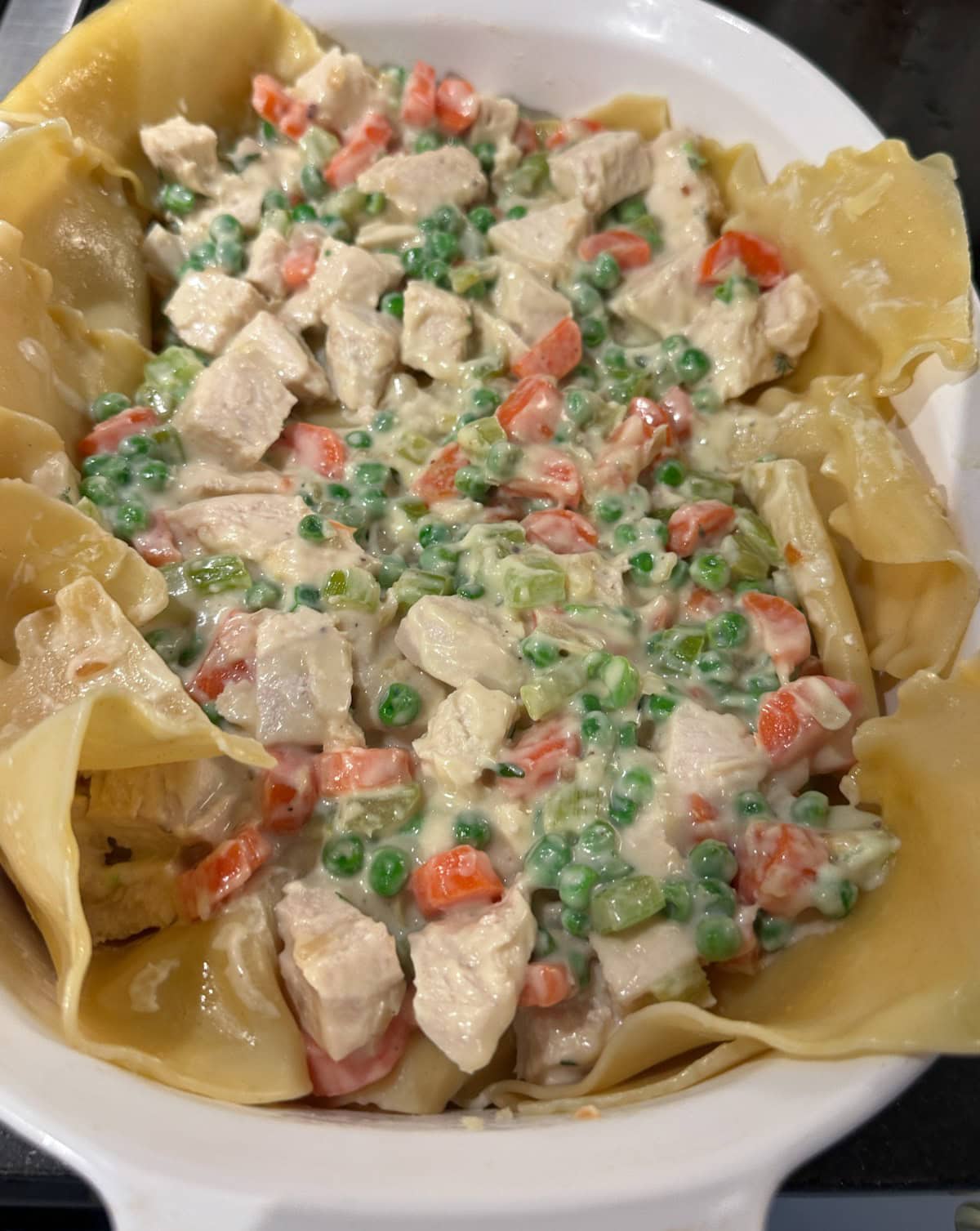 A casserole dish filled with creamy chicken pot pie ingredients. Visible are chunks of chicken, peas, and carrots in a creamy sauce, surrounded by uncooked pastry dough.