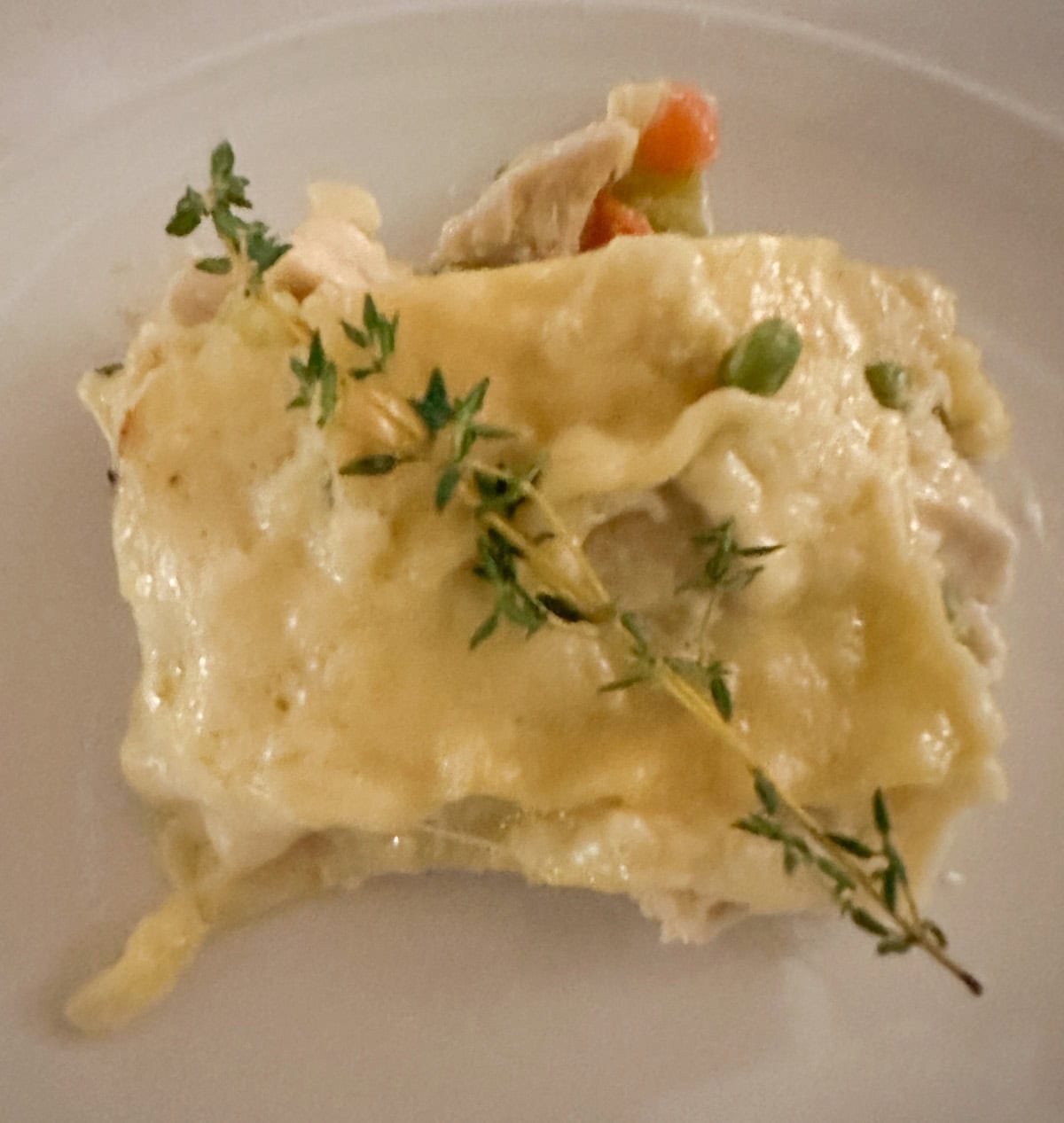Close-up of a slice of creamy lasagna on a plate, topped with a sprig of fresh thyme. The dish includes visible layers with cheese, pasta, chicken, and vegetables like carrots and peas.