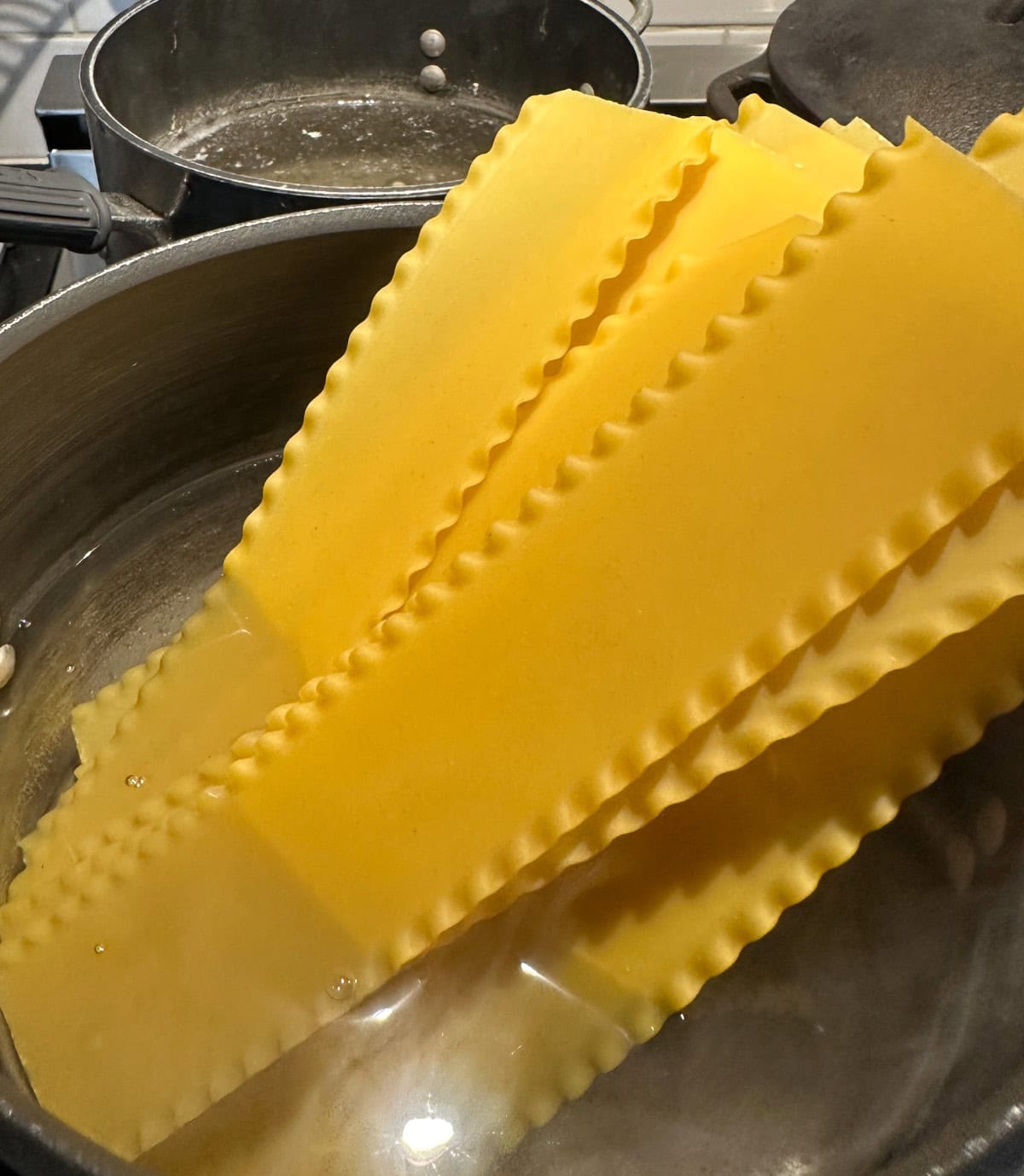 Lasagna noodles being boiled in a pot of water on a stove, with another pot visible in the background. The noodles are partially submerged and softening in the hot water.