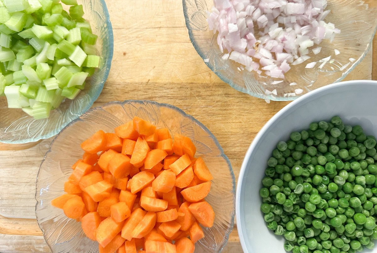 Four bowls of chopped vegetables on a wooden surface: celery, onions, carrots, and peas. The vegetables are neatly diced and vibrant in color.
