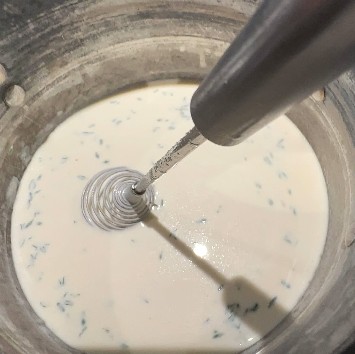 A metal whisk is blending a creamy liquid mixture with green herbs, inside a metal bowl. The liquid appears smooth and speckled with small bits of herbs, creating a subtle texture.
