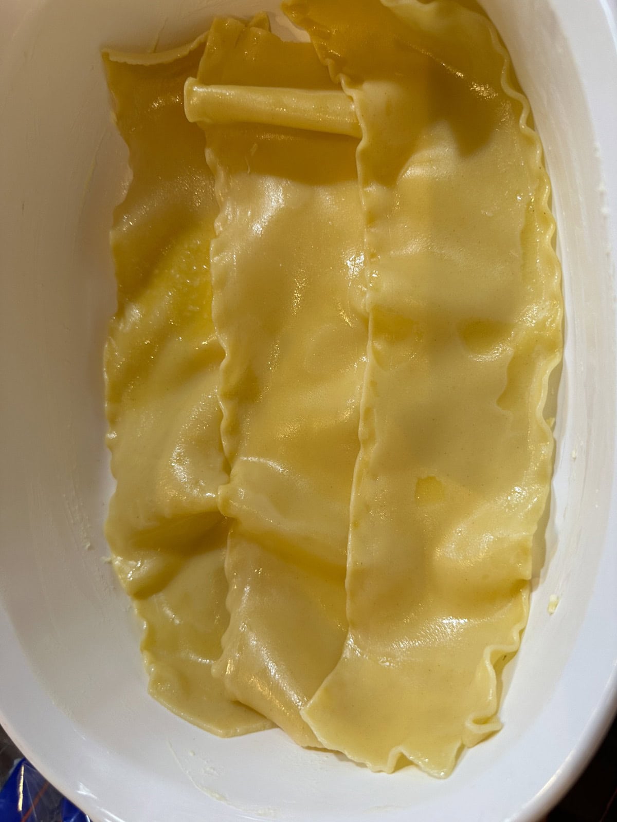 Uncooked lasagna sheets are placed flat in a white rectangular dish, with visible ridges along the edges. The sheets have a light yellow color and a glossy surface, likely from being pre-boiled.