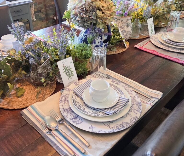 A beautifully set dining table features elegant floral-patterned plates, striped napkins, and white teacups on linen placemats. Centerpieces include fresh herbs, flowers in jars, and a blue floral vase, crafting an inviting ambiance perfect for sharing a delightful meal or hosting a cozy podcast discussion.