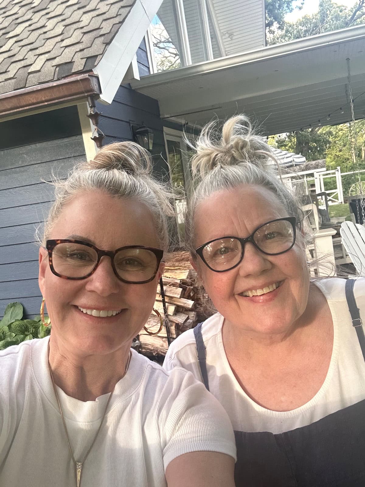Two women with light hair in messy buns and glasses smile at the camera outdoors during their Saturday shopping. They stand in front of a house with dark siding, white trim, greenery, and stacked wood behind them.