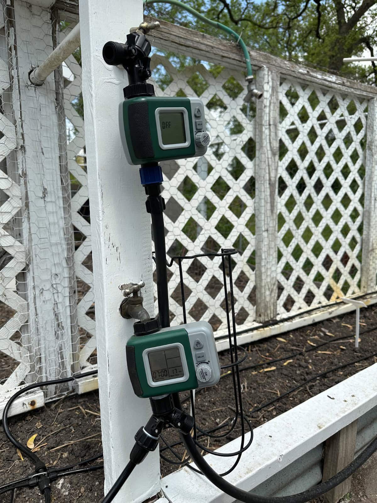 Two digital water timers are mounted on a white wooden post near a garden bed with lattice fencing in the background. Both timers are attached to garden hoses and irrigation tubing.
